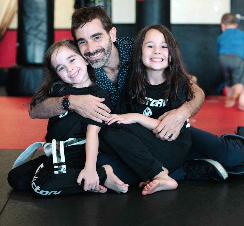A dad hugs his two grinning daughters on the mat, all three in black Victory shirts