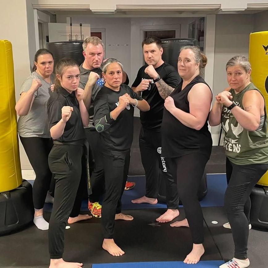 An adult Krav Maga class stands together in fighting stance in front of the heavy bags