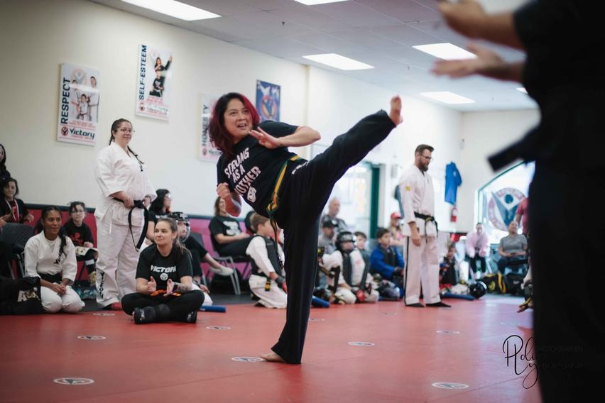 An adult black belt in a white Victory uniform throws a high front kick toward a teenage training partner