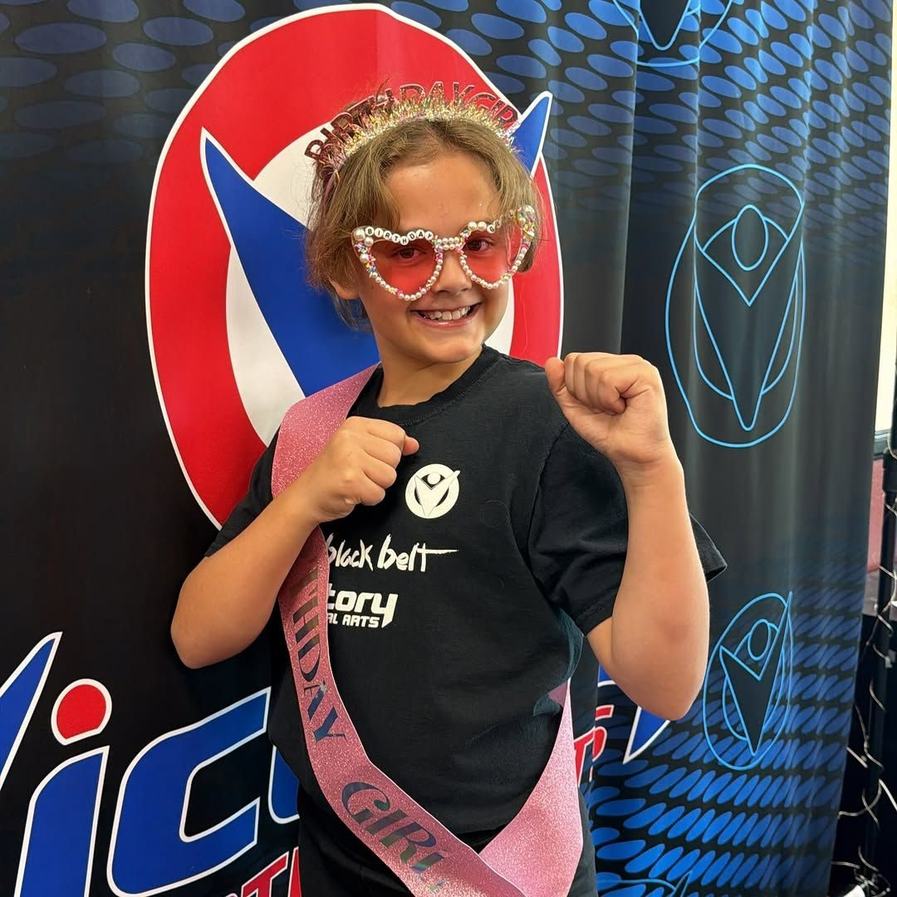 A birthday girl in a crown and sash flexes in front of the Victory backdrop